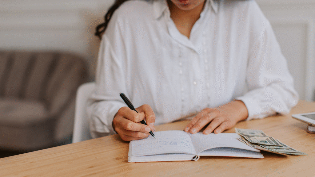 Person writing in a notebook with cash on the table, budgeting and saving money for a future home purchase.