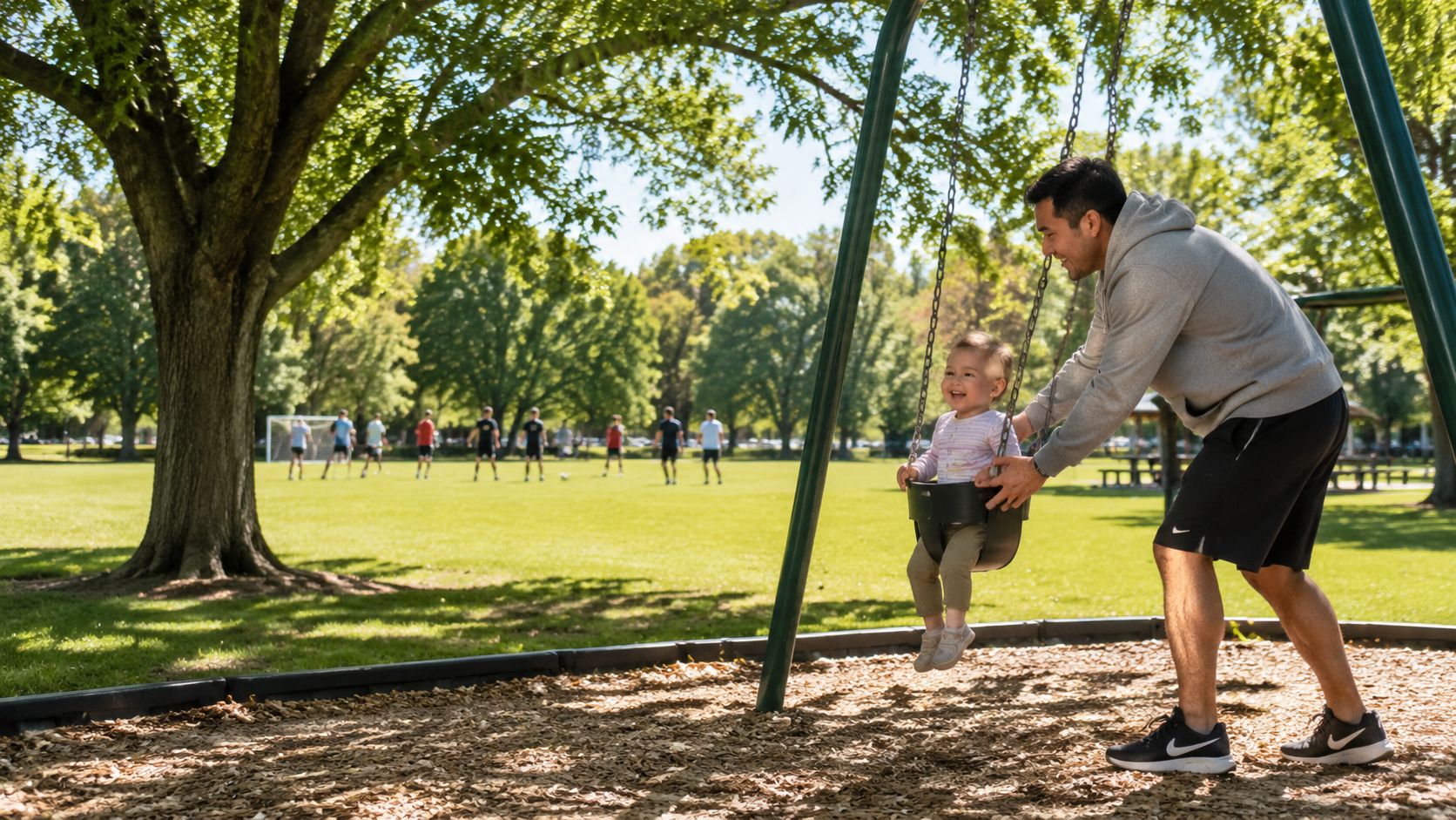 A father pushes his smiling toddler daughter on a swing set at a sunny public park.